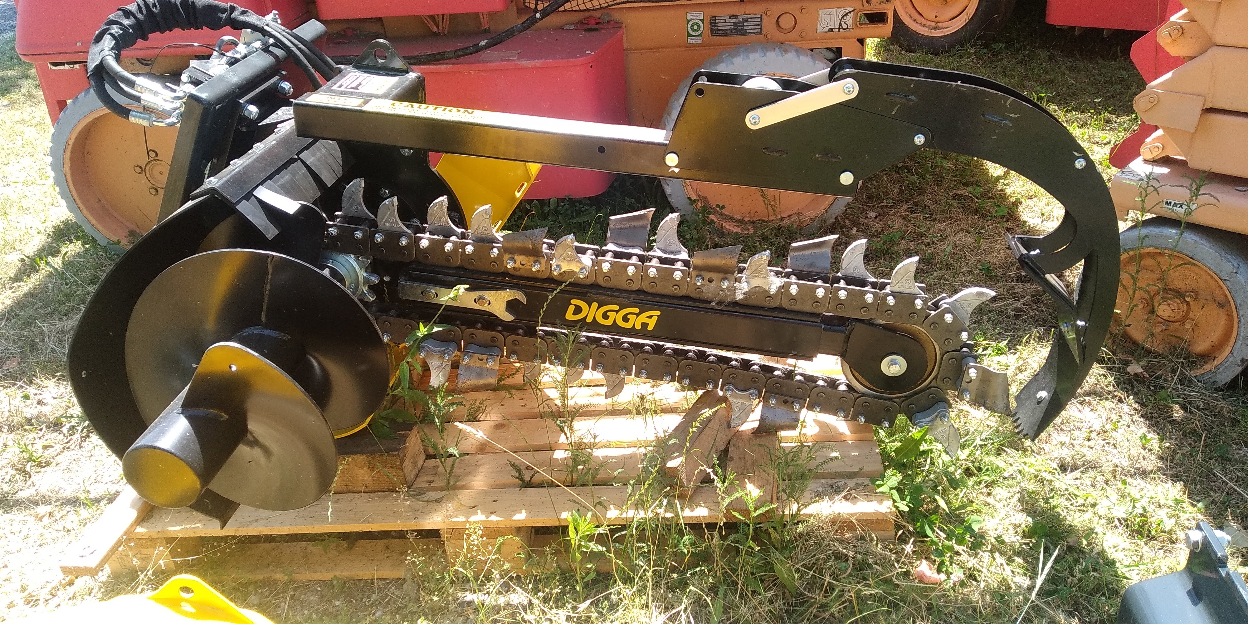 Heavy-duty black Digga trencher attachment with chain and spade resting on wooden pallets outdoors.